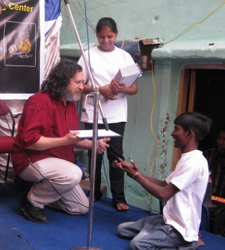 A photo of Richard Stallman kneeling down to reach a handicapped student sitting on the floor.